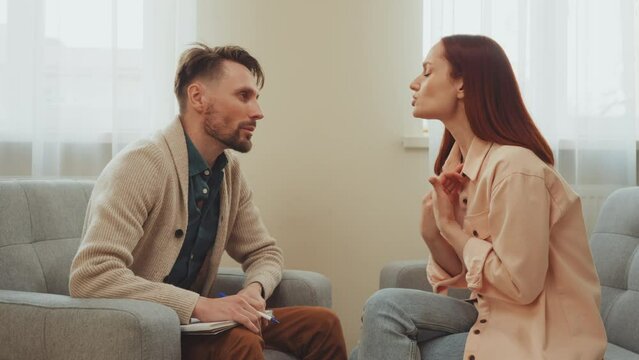 Distressed woman in emotional therapy session with male psychologist, seated on gray sofas. The scene captures moment of support and understanding in calming, professional environment