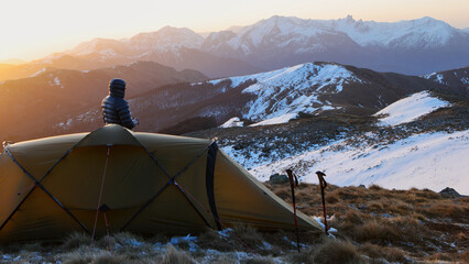 A hiker stands beside a tent, admiring a sunrise over snow-covered mountains. Walking poles are...