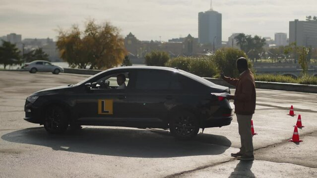 Full shot of male African American instructor supervising young woman studying forward and reverse driving at outdoor urban parking area