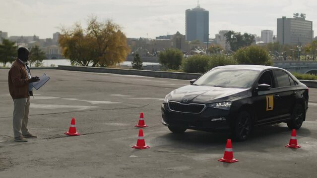 Full shot of angry African American driving instructor evaluating students parking test on learners vehicle with L plate, shouting for knocking over orange cone