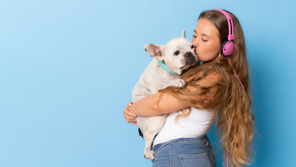 Young smiling woman hugging her dog wearing headphones standing isolated over blue background