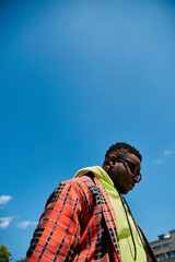 Fototapeta premium Handsome African American man in fashionable plaid jacket standing under a clear blue sky.