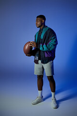 Fototapeta premium A fashionable young African American man holding a football in front of a blue background.