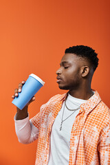 Handsome African American man sips from cup against vibrant orange backdrop.