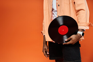 Fashionable young African American man in stylish attire holding a vinyl record against an orange...