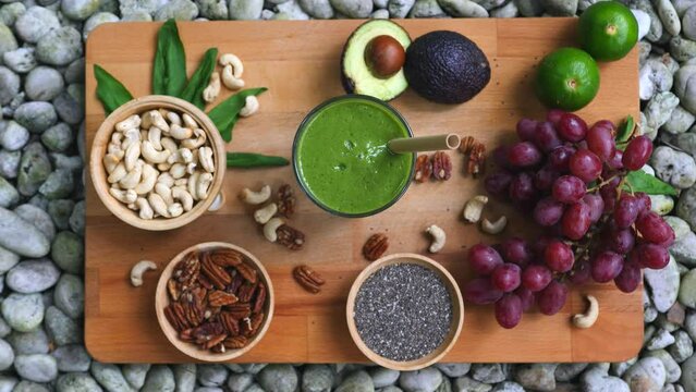 Flat lay of healthy green fruits, vegetables, nuts and seeds on wooden board for healthy eating. Woman hand places glass of healthy smoothie with straw. Healthy morning breakfast with green smoothie
