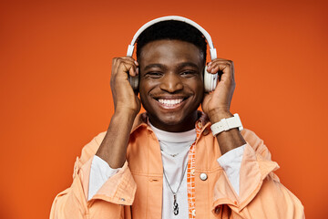 Handsome African American man in stylish attire, smiling while wearing headphones on orange...