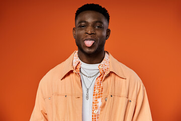 A fashionable, young African American man sticks out his tongue while wearing a stylish orange jacket.