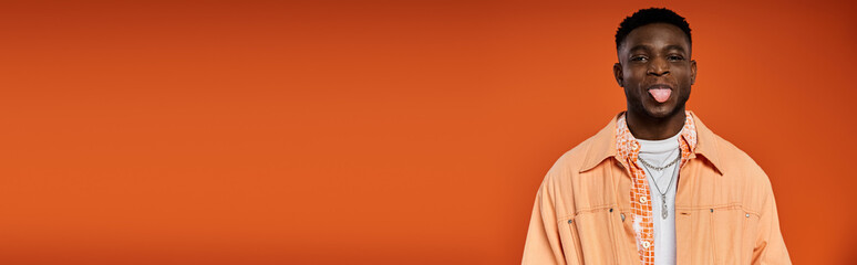 Young African American man in stylish attire sticks out his tongue against orange background.