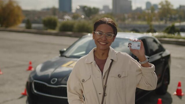 Portrait of young cheerful African American woman wearing glasses, smiling and showing drivers license at camera, sunny urban landscape in background