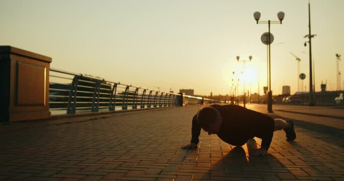 Young athletic man training outside doing push-ups on a city street at sunset. Outdoor workout exercises. Active healthy lifestyle, wellbeing and body care. Slow motion wide shot.
