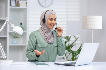 Smiling woman in a hijab using a headset on a video call, working on a laptop in a modern home...