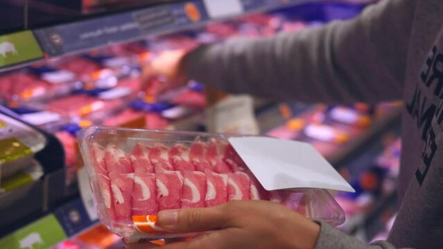 Close-up hand holding packaging fresh meat, choosing through bacon on shelf. Right packaging is key to food safety and freshness. Buying bright and catchy packaging of meat slices in supermarket.