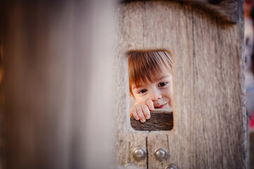 Fototapeta premium A cheerful toddler in overalls peeks through an opening in a wooden playground structure, smiling brightly, with an autumnal background.