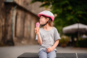 A young girl enjoys a pink ice cream cone while sitting outside, wearing a bike helmet and casual...