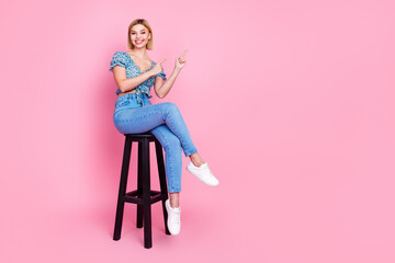 Full size photo of pretty young girl sit stool indicate fingers empty space wear top isolated on pink color background