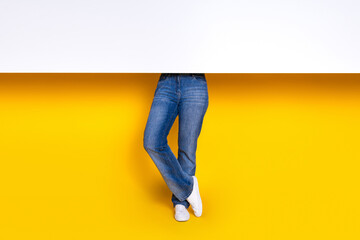 Cropped photo of confident woman legs dressed jeans trousers sneakers hold white placard empty space isolated on yellow color background