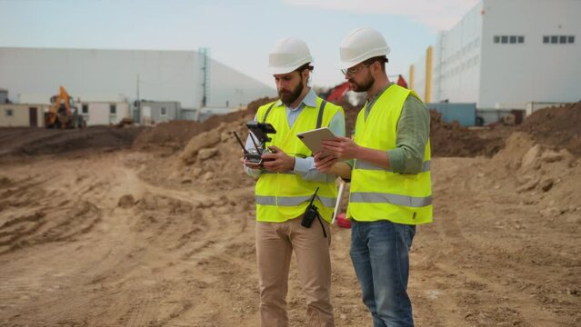 Two builders in bright yellow vests holding tablet and smartphone while controlling flying drone observing construction site. Topographers on background. Outdoors