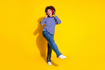 Full length photo of funky lovely woman dressed striped shirt jeans dancing touching headphones isolated on yellow color background