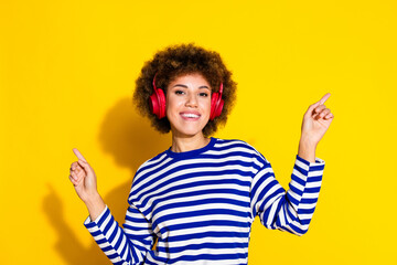 Portrait of toothy beaming girl with perming coiffure dressed pullover listen music in headphones isolated on yellow color background