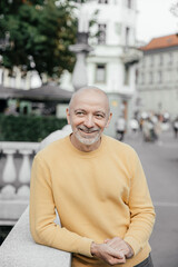 Cheerful bald senior man in a yellow sweater, leaning on a stone balustrade in an urban...