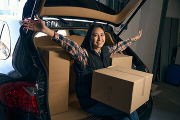 Smiling lady is sitting in the trunk of a car with boxes, raising her hands up