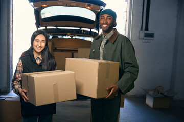 Smiling man and woman are unloading things from the trunk of a car