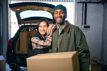 Smiling woman hugs a man holding a box in his hands near a car
