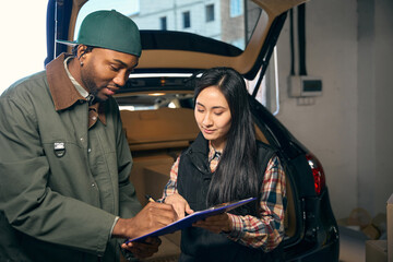 Couple filling out folder, standing against background of car with open trunk