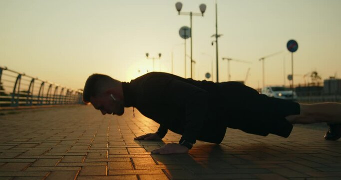 Young athletic man training outside doing push-ups on a city street at sunset. Outdoor workout exercises. Active healthy lifestyle, wellbeing and body care. Slow motion medium shot.
