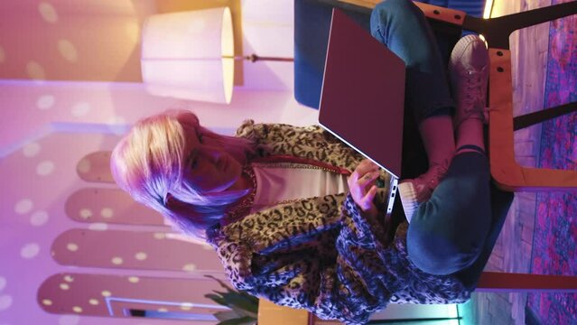 Disco neon stroboscopes illuminate room. Beautiful fashionable glamorous young woman sitting on chair using modern laptop to check bank account. Stylish girl works at home during vacation.