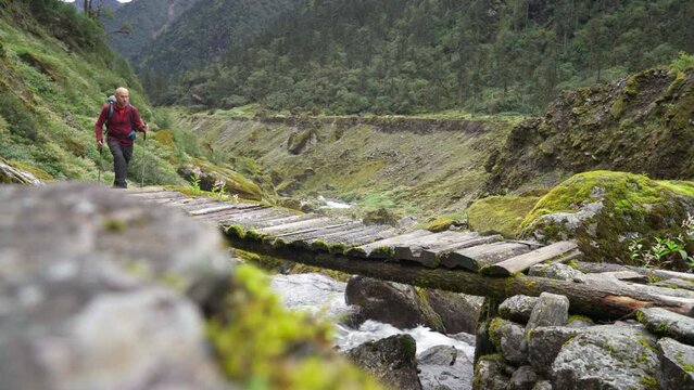 Man with backpack and trekking poles crossing mountain creek wooden bridge during Makalu Barun National Park trek in Nepal. Mountain hiking and active people concept image.