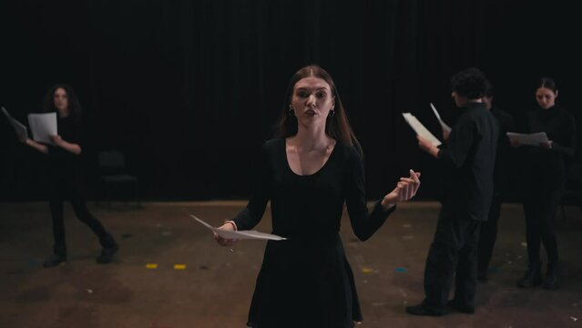 A confident young girl with glowing hair color in a black suit comes closer and reads out her text from a piece of paper while playing her role in the theater impression of emotions on stage during a