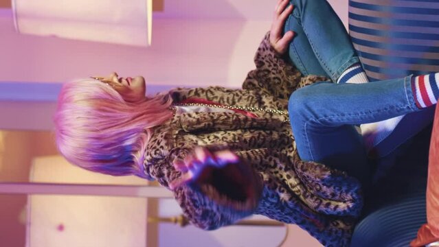 Pretty young creative beautiful woman sitting and resting on soft vintage sofa. Trendy cool magnificent girl in sunglasses dancing, hanging out in retro studio. Disco stroboscopes illuminate room.