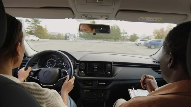 Back view of young female biracial student at driving school navigating left-handed car under supervision of mature instructor holding clipboard