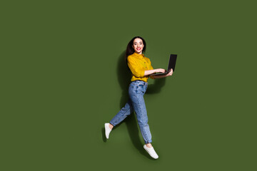 Full length photo of adorable lovely girl wear yellow shirt typing modern gadget jumping high isolated green color background