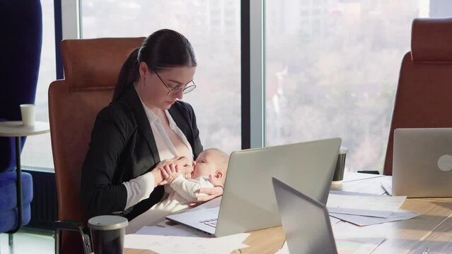 A confident brunette girl in round glasses in a business uniform sits at a table in front of a laptop in the office and breastfeeds her baby without being distracted from work in a modern office