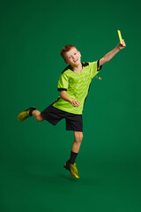 Little boy, kid referee showing yellow card and joyfully jumping against vibrant green background....