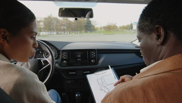 Back view of mature male African American instructor sitting in passenger seat explaining parallel parking technique to female student in her twenties learning to drive