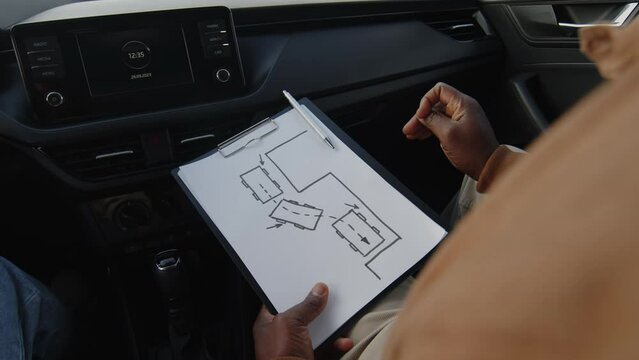 Over-shoulder close-up shot of hands of male African American instructor sitting inside car teaching offscreen student of driving school, explaining parallel parking rules by showing scheme