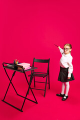 Fototapeta premium Child in glasses and formal attire, as teacher holding pointer stick and pointing on imaginative writing board against vibrant pink background. Concept of dreams, future career, games, fantasy. Ad
