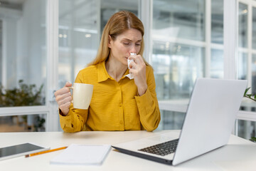 A woman in a yellow shirt sits at her desk, drinking tea and blowing her nose, indicating illness...