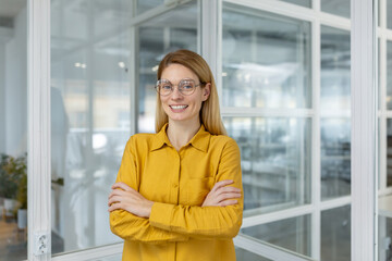 Confident businesswoman in a yellow shirt and glasses standing with arms crossed in a modern office...