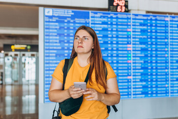 Perplexed unhappy 30s woman holding cellphone in hands, checking Trip Destination on Internet near information board. Delayed flight. Receives Shockingly Bad News, Misses Flight.