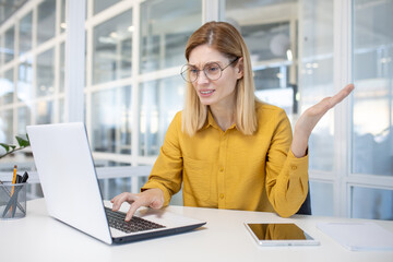 Confused professional woman with glasses using a laptop in a modern office setting, showing uncertainty and contemplation.