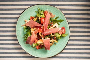 Bright salad with spinach, mushrooms, slices of prosciutto and tomatoes served on a white plate in...