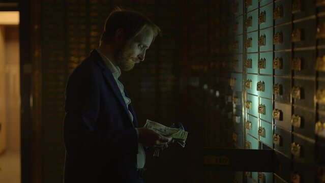 A man is inside a dimly lit vault handling money and looking at safety deposit boxes