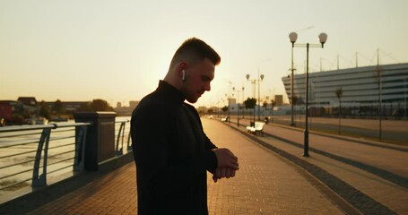 Young active athlete using smartwatch before starting jogging at sunset in the city. Medium slow motion shot from behind. Using wearable technologies in everyday life. Active healthy lifestyle. - Powered by Adobe
