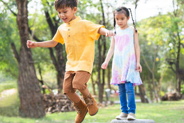 Group of multiracial children in line to jumping from stone chair to another one chair outdoors in summer park. Adorable kids have fun playing together in garden. Selective focus on little boy