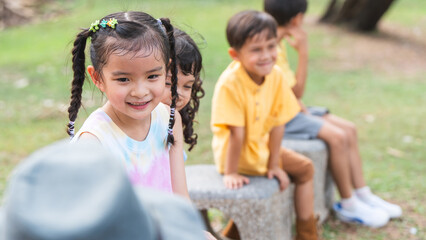 Group of multiracial children smiling, sitting at stone chair, playing together outdoors in summer park. Adorable kids have fun playing in playground. Selective focus on Asian girl